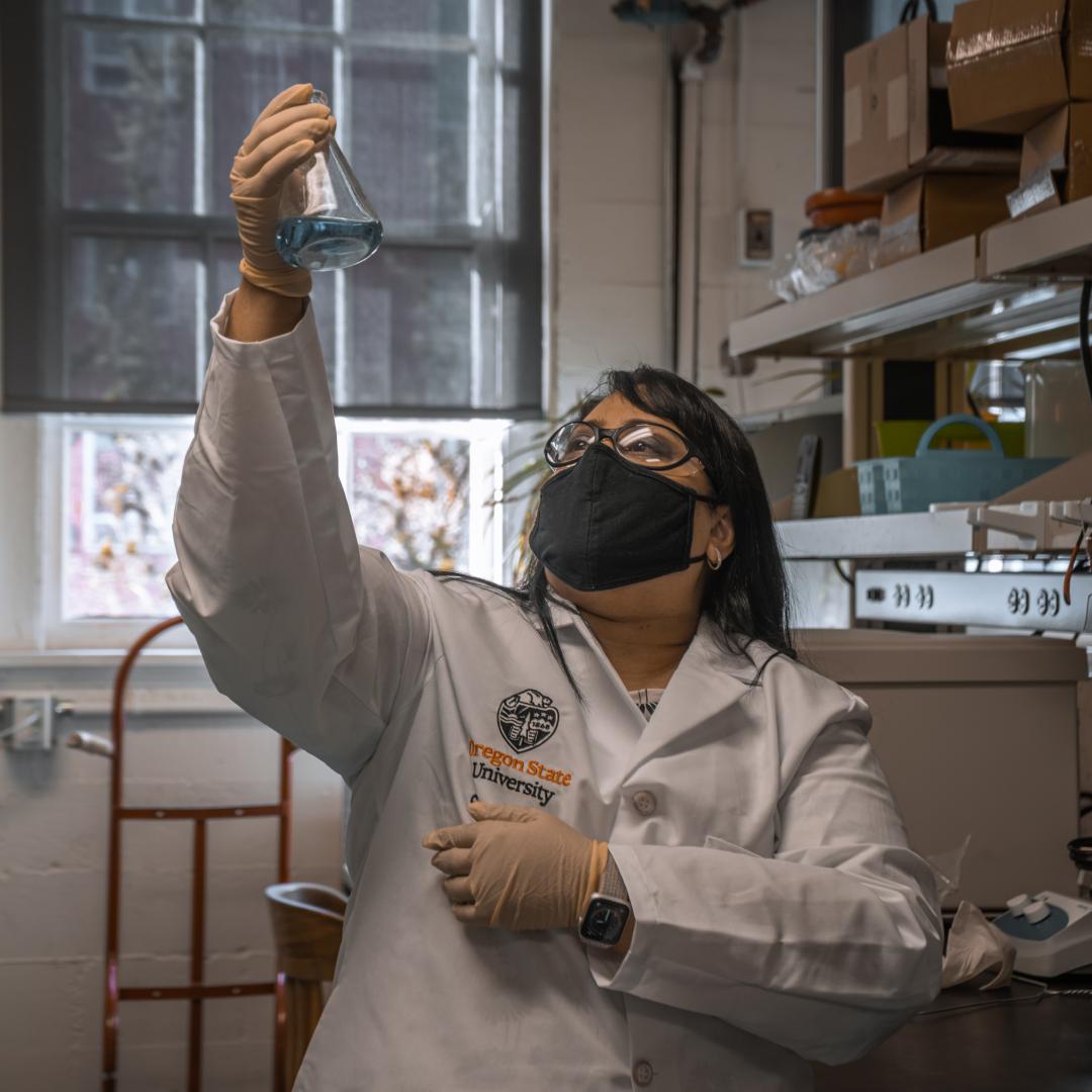 Female scientist in white lab coat looks up at a beaker with blue liquid inside it.