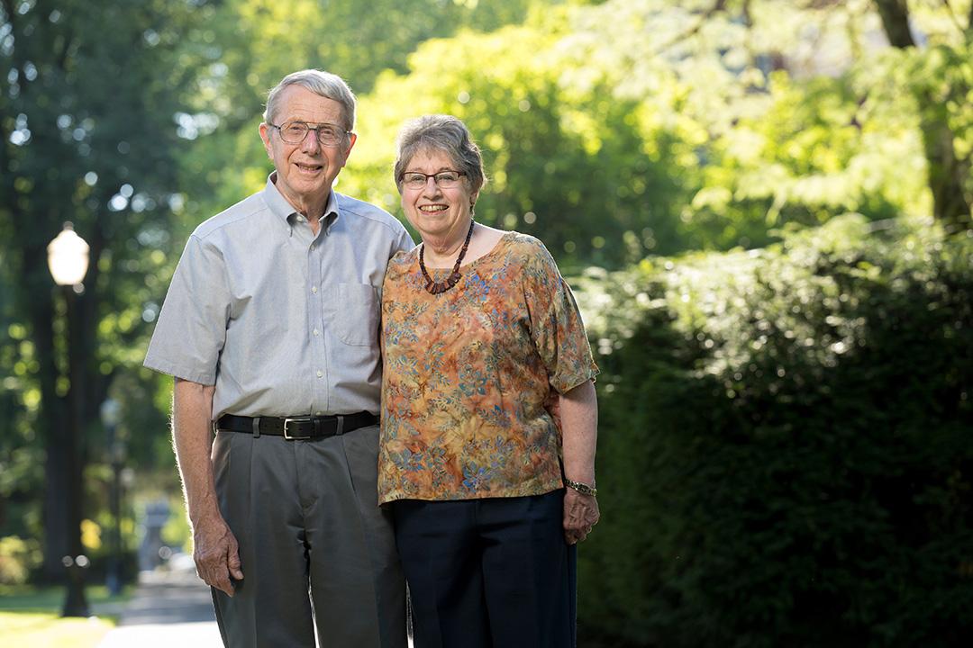A man and woman smile together in front of a background of trees.