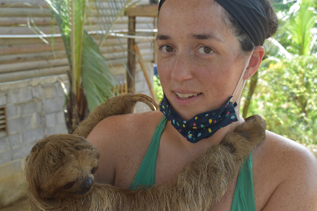 A woman in a green tank top holds a sloth in her arms, surrounded by tropical foliage.