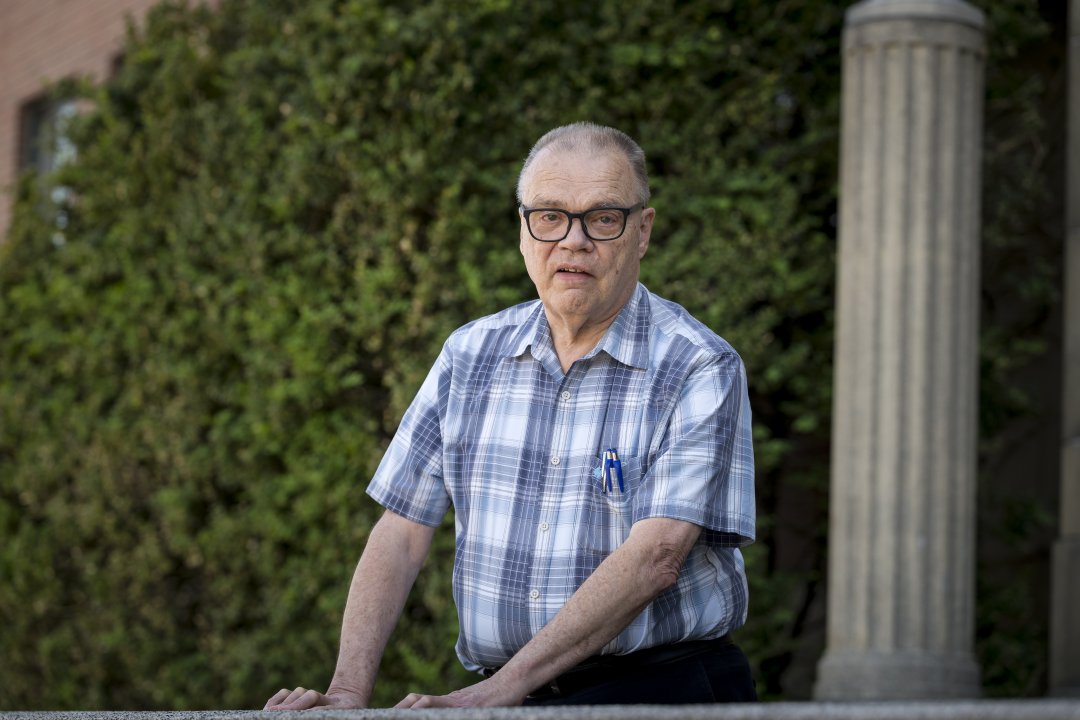 A man in a plaid shirt stands in front of a green background.