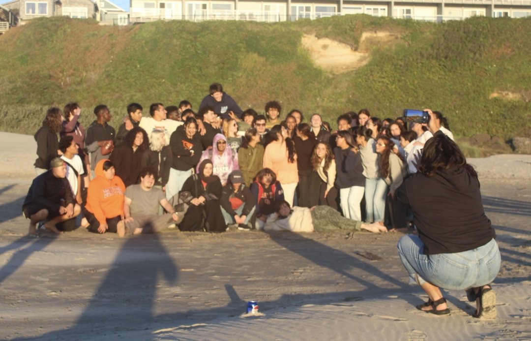 Under a setting sun, a large group of students huddle on a beach while a woman in the foreground kneels to take their picture.