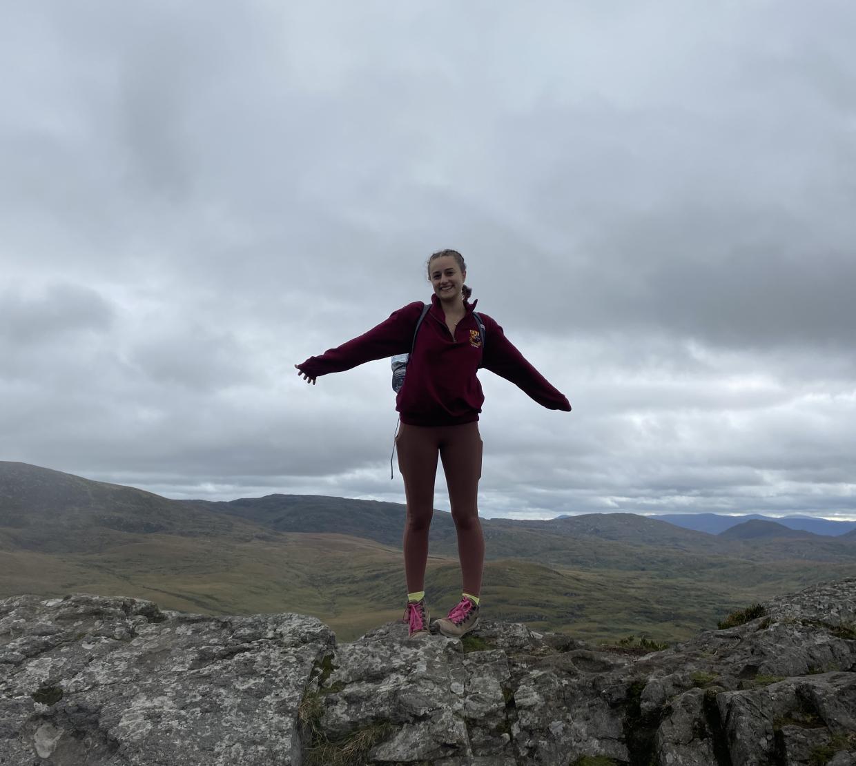 Amelia Noall standing at the top of Torc Mountain in Ireland, overlooking a vast field.