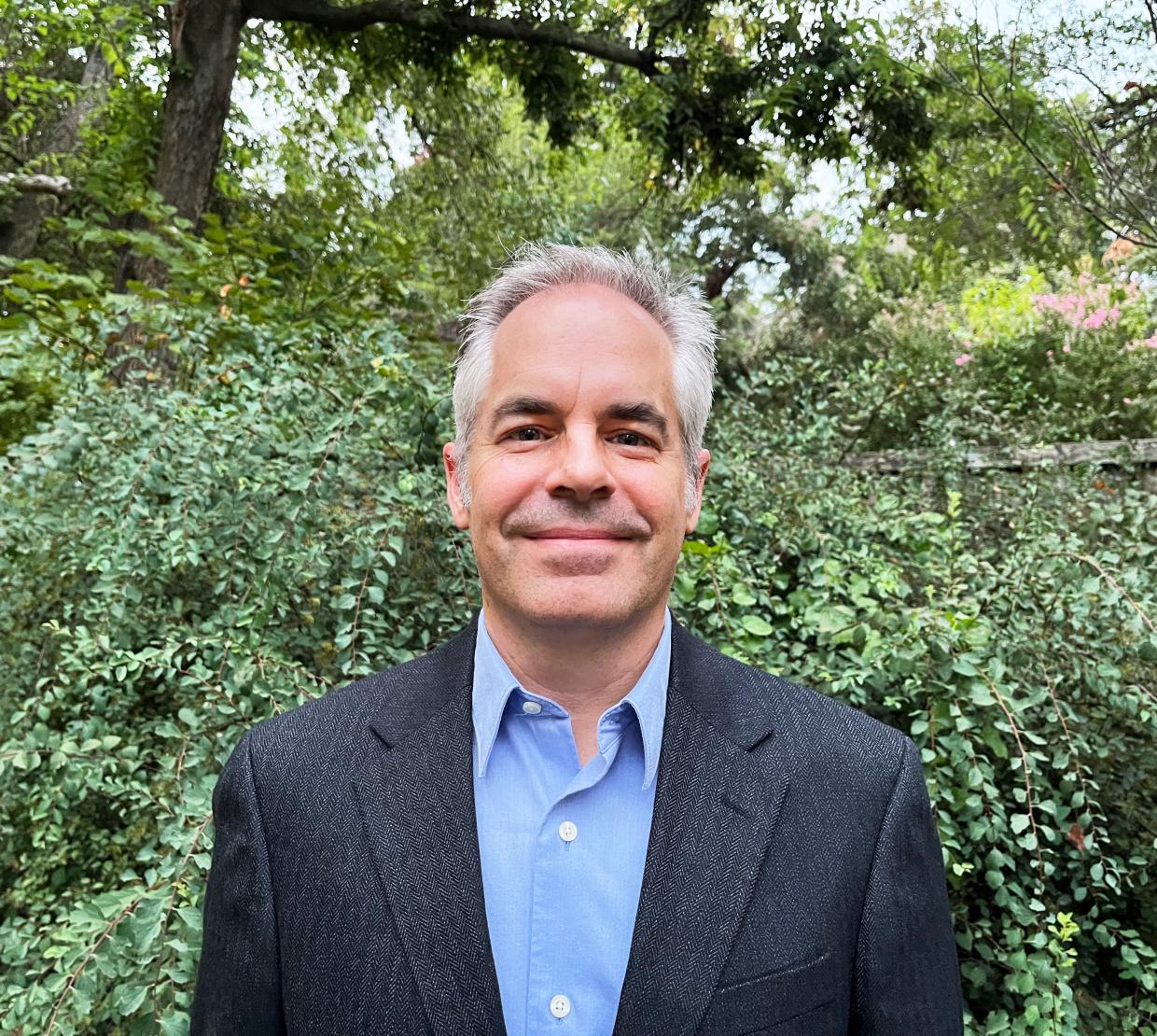 Professor Jon Kujawa stands in a suit coat and smiles in his headshot outdoors surrounded by greenery. 