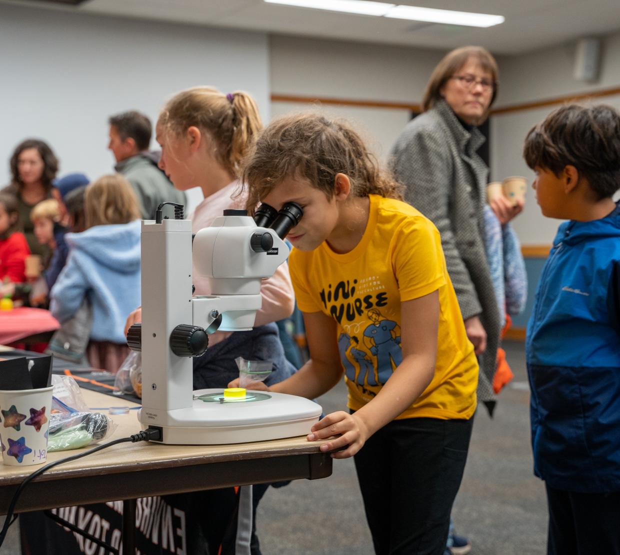 A young student peers into a microscope surrounded by the bustle of Discovery Days.