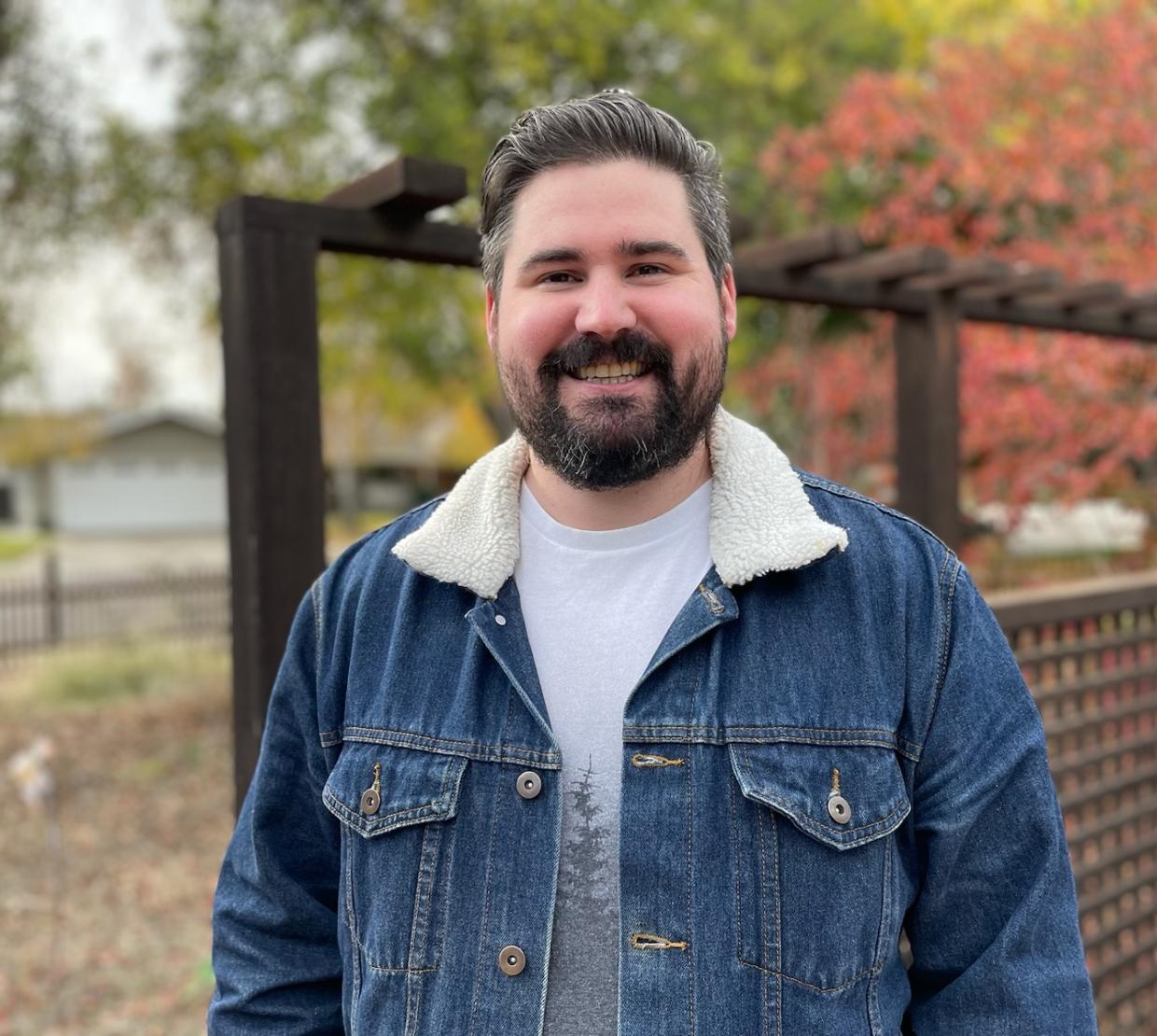 A man with a beard stands in a denim jacket with autumn colored trees in the background.