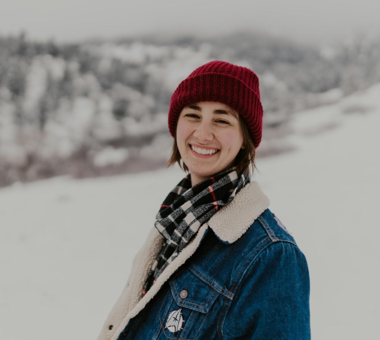 Megan Tucker smiles widely, her scarlet beanie a striking contrast to the snow-covered mountaintops that rise behind her.