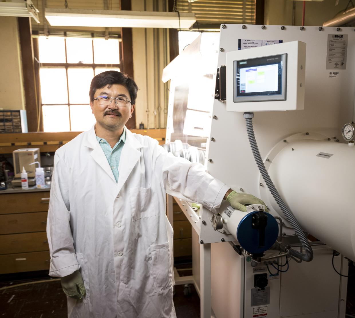 A man in a lab coat stands in front of a white machine used for battery science.