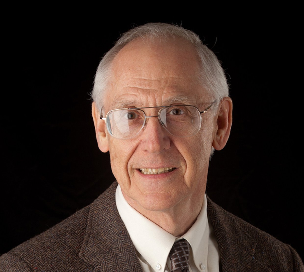 A man's headshot against a black background. The man has gray hair and wears glasses with a brown suit jacket and tie.