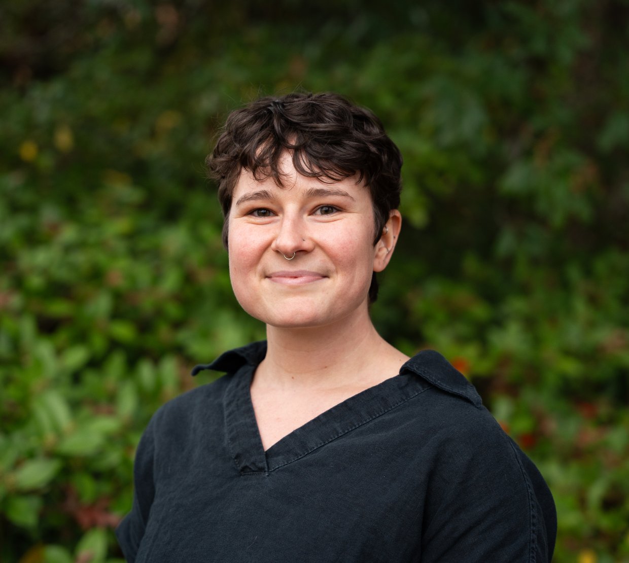 Sophia Bailey smiles in front of Oregon greenery. She wears a black button-up shirt and has short cropped, curly, dark hair. 