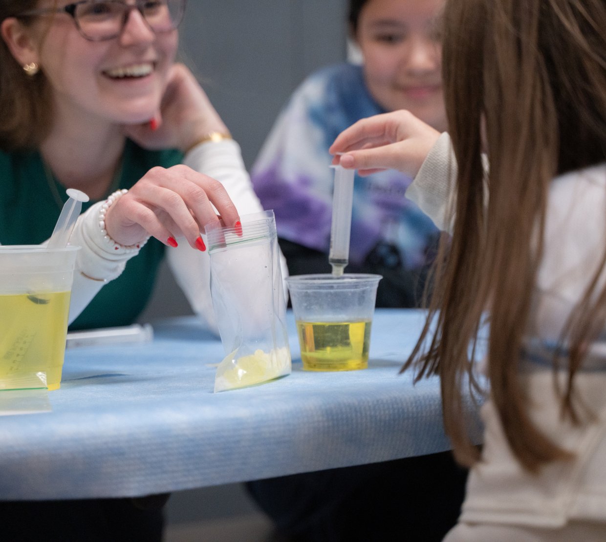 An OSU student smiles widely at the child in front of them. She holds a baggie of yellow liquid that the child is using a syringe to extract and place into a cup in front of her.