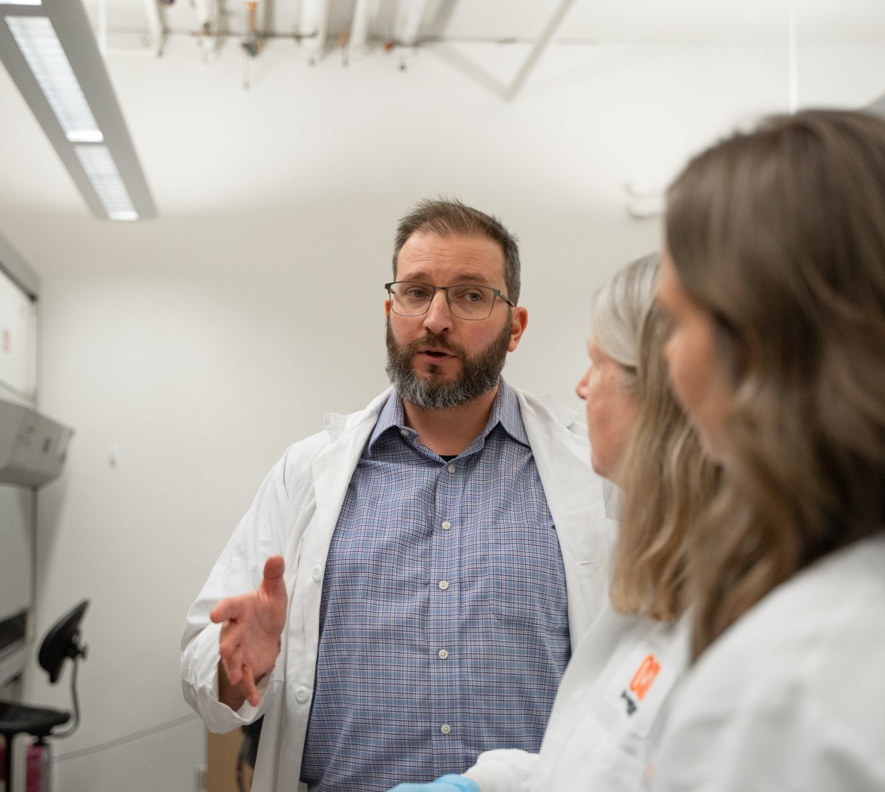 A bearded man wearing glasses and a button-up shirt beneath a lab coat speaks to two other scientists wearing lab coats toward the camera.