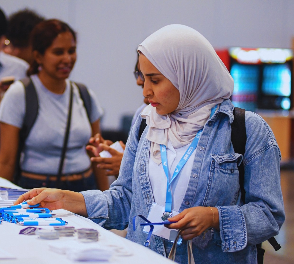 Woman wearing a hijab and a blue JSM lanyard peruses merchandise on a table with other conference attendees milling around.