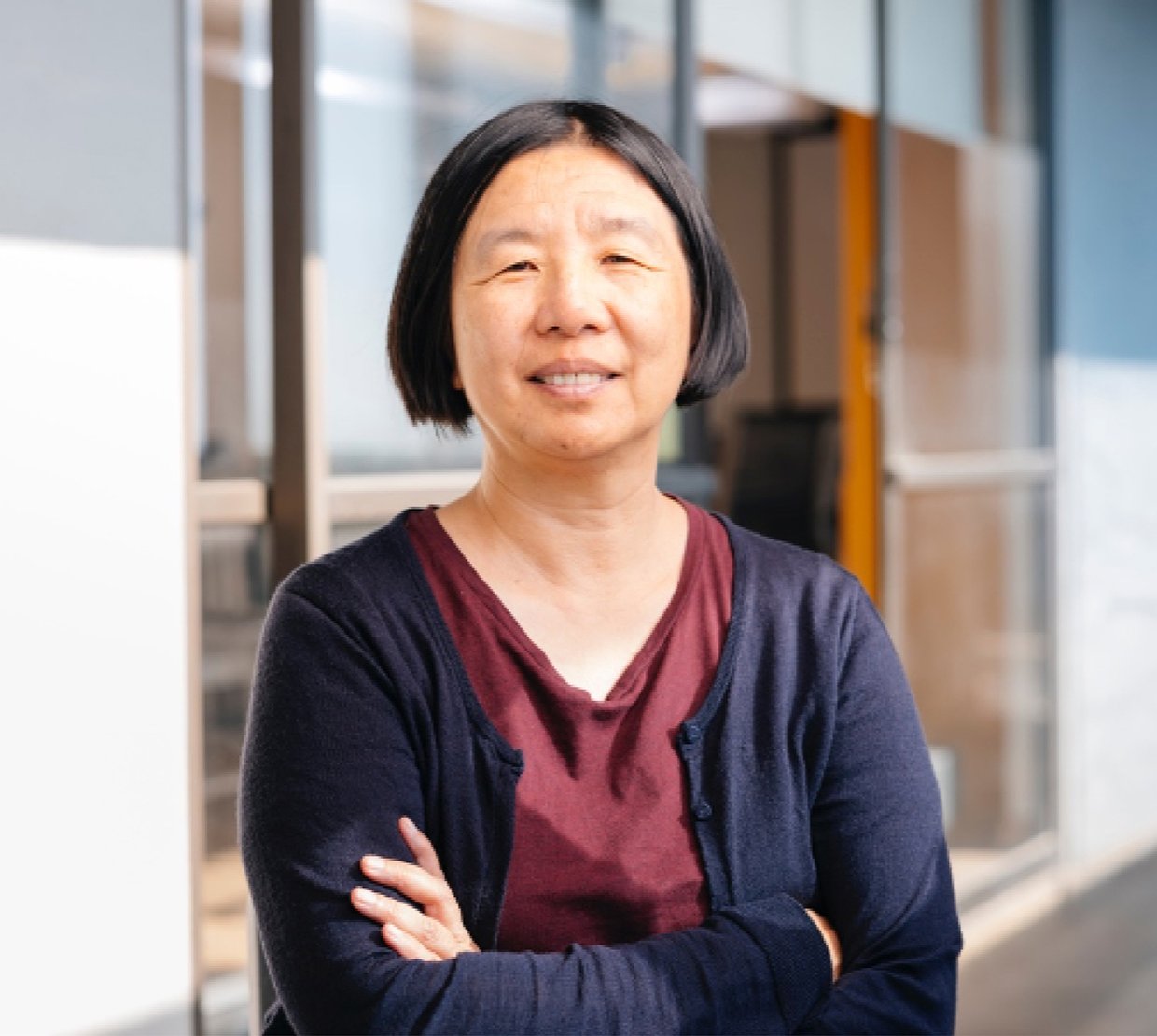Headshot of Professor Bin Yu of UC Berkeley: woman in dark blue cardigan and maroon blouse, smiling with arms crossed while standing in front of college building with large glass windows.