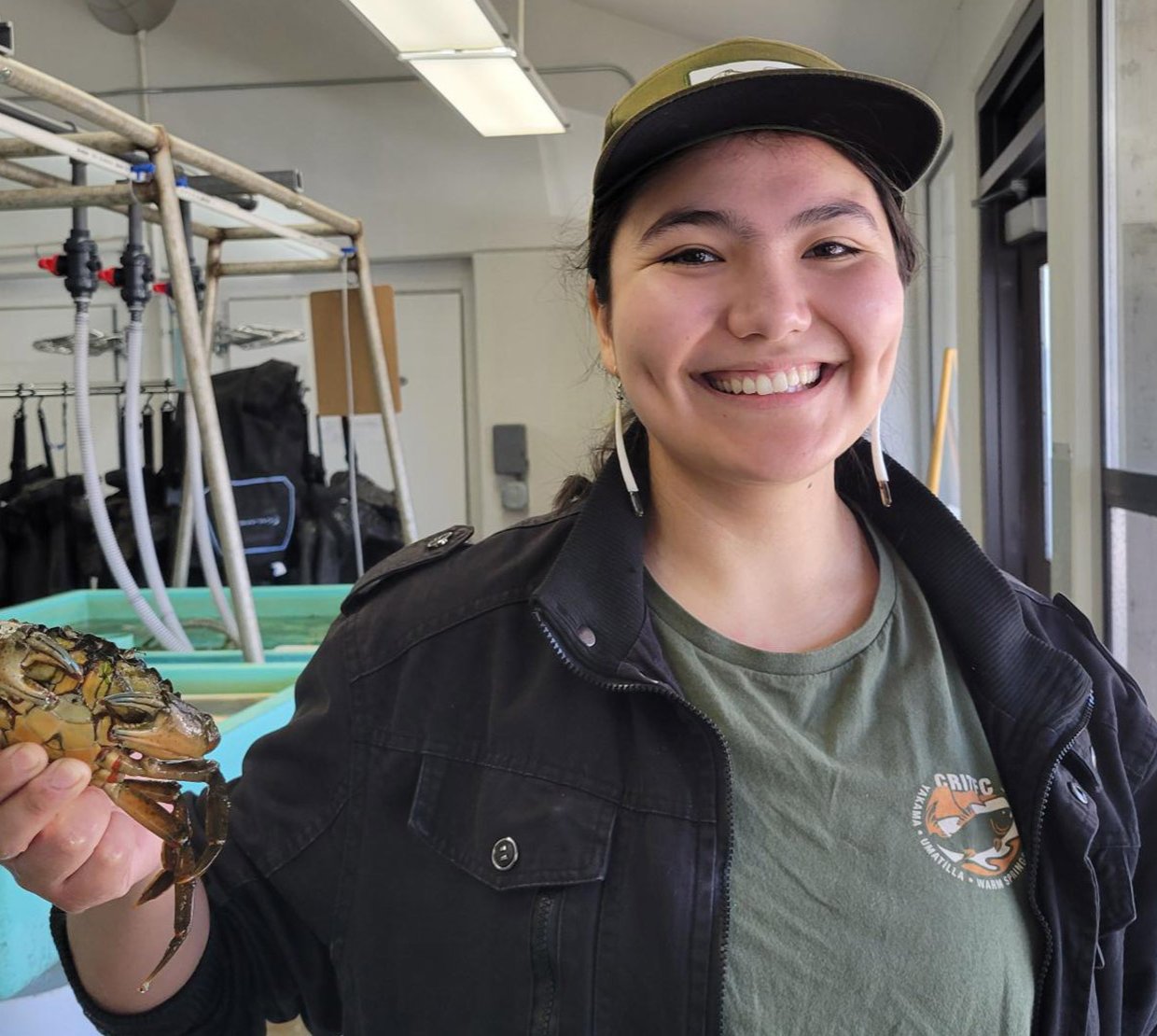 Ingram standing the Hatfield Marine Science Center wet lab, holds up a crab. She wears a CRITFIC shirt, baseball cap and beaded earrings.