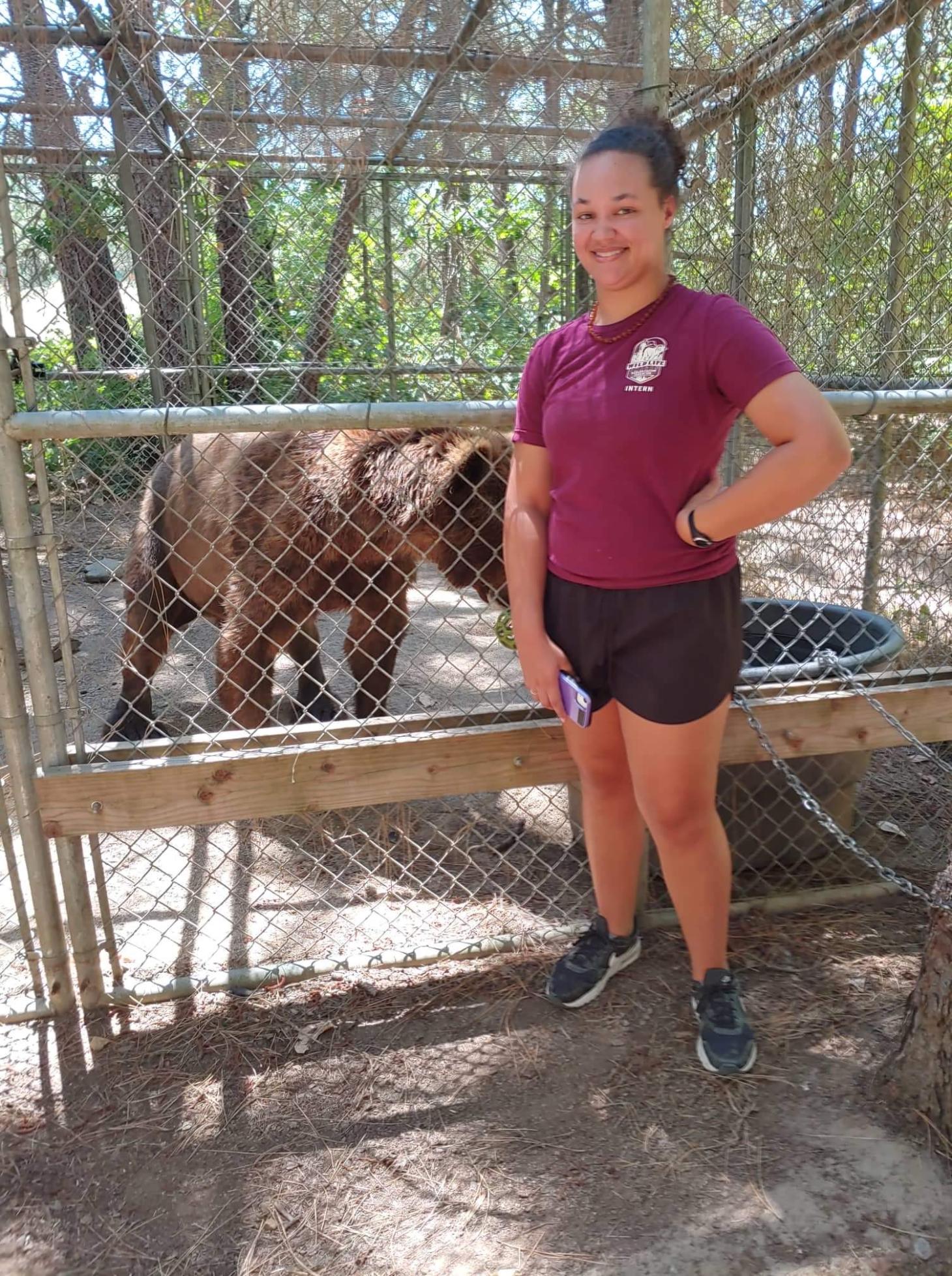 Camrie poses in front of a brown bear's enclosure at Wildlife Images Rehabilitation Center. She wears a Wildlife Images Tshirt and smiles broadly. 