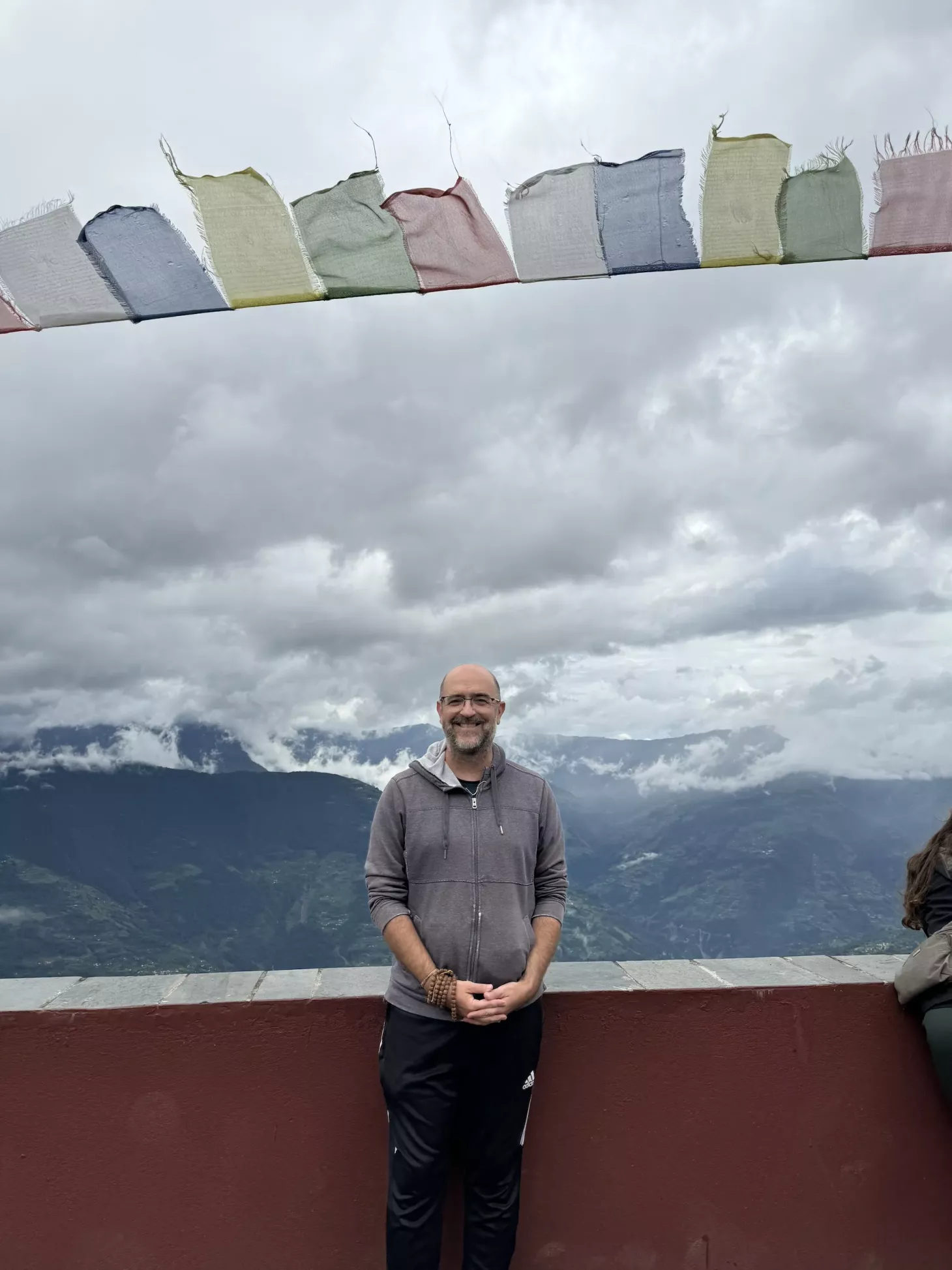 Professor Dee Denver smiles under prayer flags with misty mountains in the distance.