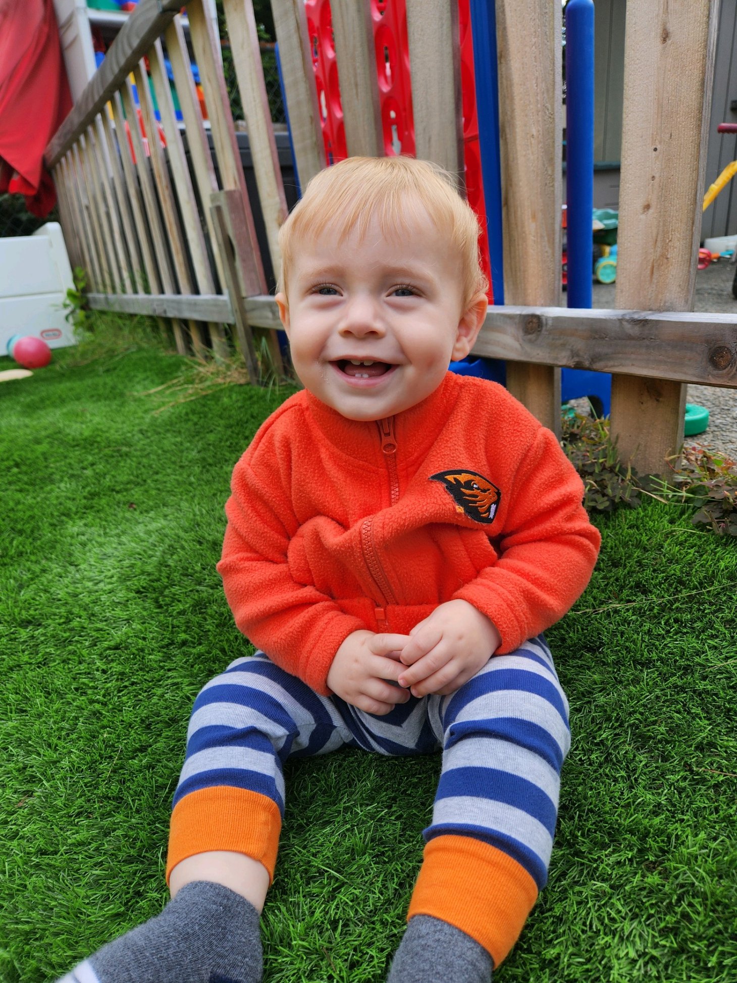 Laith Rogers, as a baby, sitting on the grass and laughing while wearing an orange Oregon State University fleece.