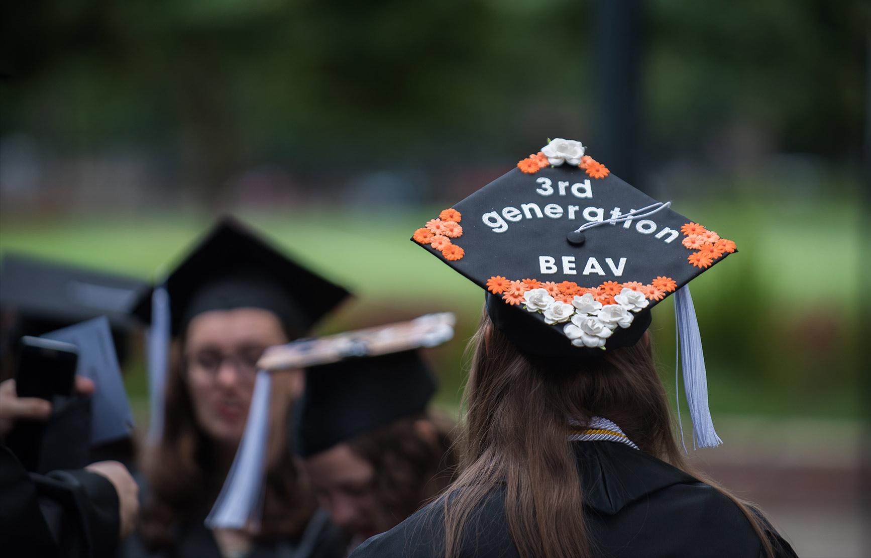 Graduating student in crowd of graduates, wearing a grad cap with "3rd generation BEAV" written on with flower decorations.