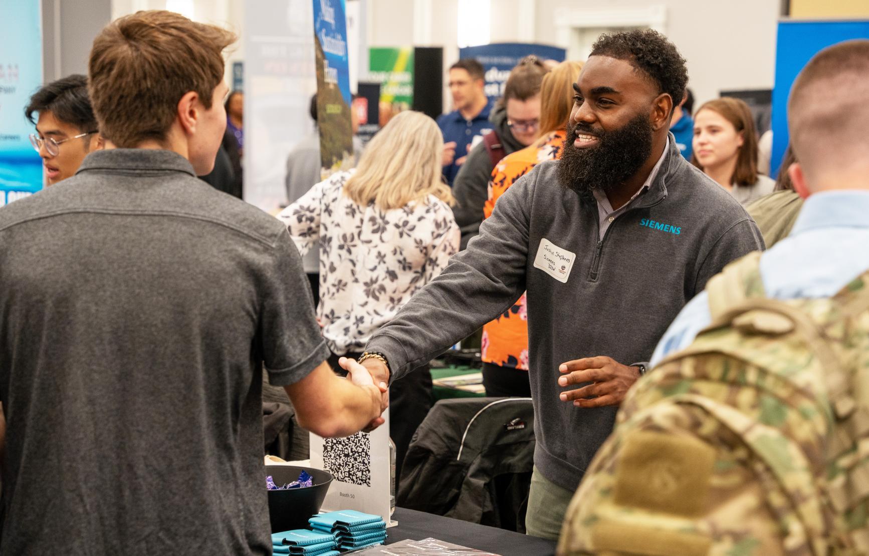 An industry professional smiles and shakes hands with a student across a table. Their shirt reads "SIEMENS."