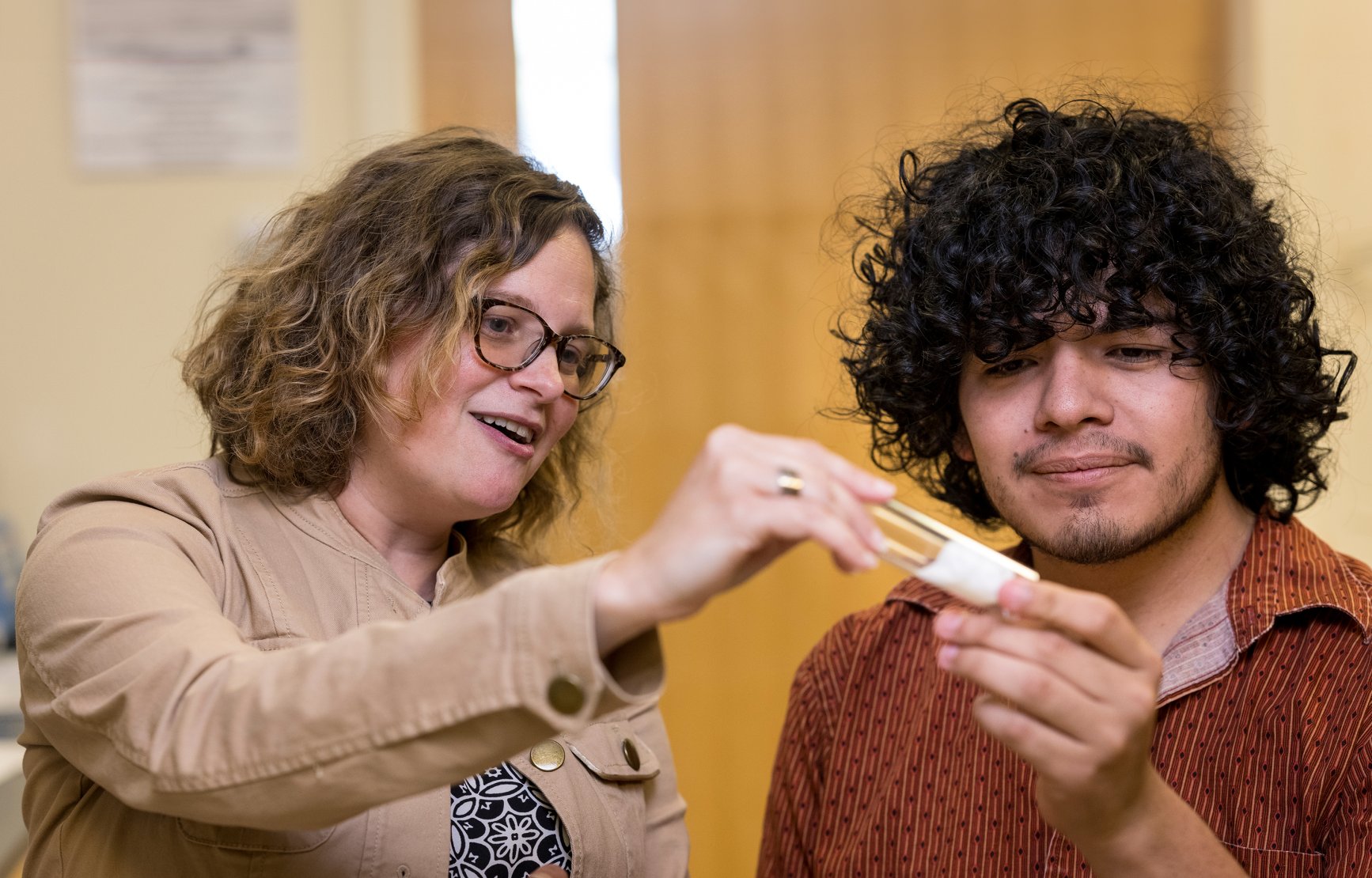 A student researcher looks at a vial of fruit flies with his smiling mentor