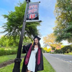 Alyssa Pratt stands in front of a pole with a sign that reads "We did it." She is wearing her graduation cap and gown.