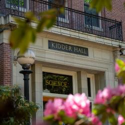 Photo of Kidder Hall from a low angle looking at a door with science written in the glass above the door, and Kidder Hall written in the stone above the glass. Blurred pink flowers appear in the foreground.