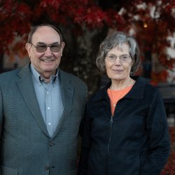 A torso-up shot of a man and woman smiling into the camera in front of a tree with red autumn leaves.