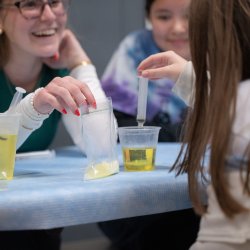 An OSU student smiles widely at the child in front of them. She holds a baggie of yellow liquid that the child is using a syringe to extract and place into a cup in front of her.