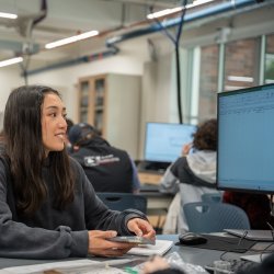 A girl in black sweatshirt works on a computer in a classroom.