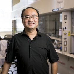 A man in a short sleeve black shirt smiles while standing in a laboratory. 