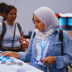 Woman wearing a hijab and a blue JSM lanyard peruses merchandise on a table with other conference attendees milling around.