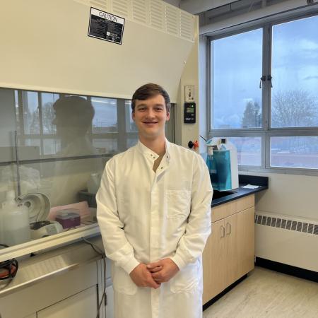 Kelly Shannon dons a white lab coat in an OSU laboratory, equipment lining the wall on his right and a window framing trees outside on his left.