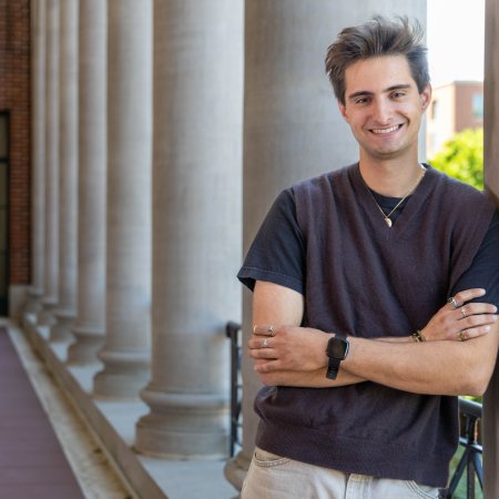 Physics student Diego Menendez leans on a large white column at Memorial Union Hall at Oregon State University. He has his arms crossed in front of him and he smiles at the camera. The hall behind him stretches on with a series of large white columns. 