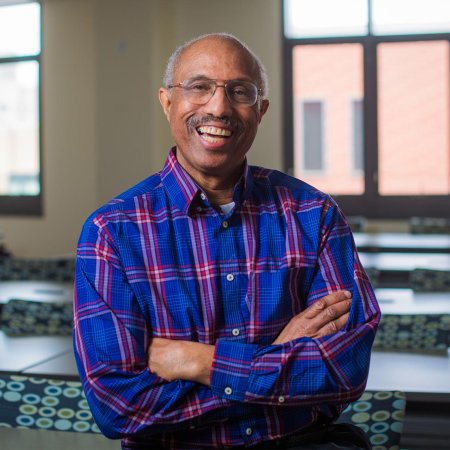 Portrait of Dr. Nathanial Whitaker: An African American male professor sitting in front of his classroom, folding his arms and smiling. 