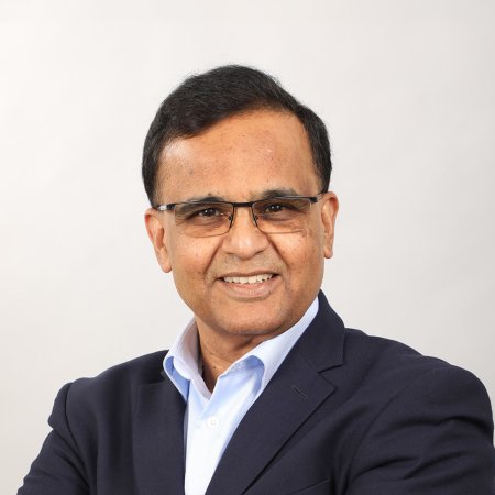 A man in a dark blazer and rectangular glasses smiles against a gray background, his short, dark hair brushed to the side.