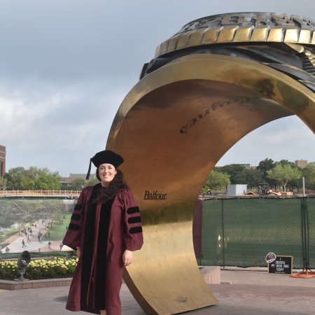 A woman wearing a deep red graduation gown stands on a college campus in front of a large golden ring statue.