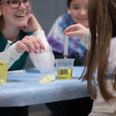 An OSU student smiles widely at the child in front of them. She holds a baggie of yellow liquid that the child is using a syringe to extract and place into a cup in front of her.