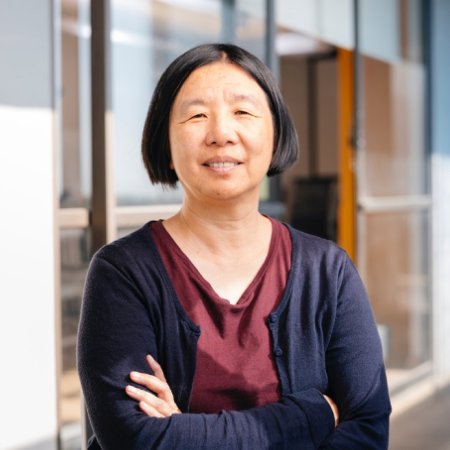 Headshot of Professor Bin Yu of UC Berkeley: woman in dark blue cardigan and maroon blouse, smiling with arms crossed while standing in front of college building with large glass windows.