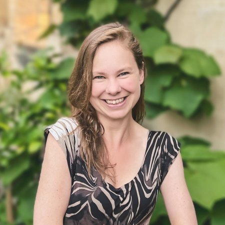 Headshot of Dr. Laura P. Schaposnik: A woman smiling with her hands folded in front of her, sitting outside a house of stone walls and healthy plants.