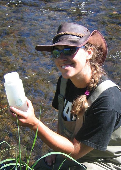 Julie Alexander equating in a river while holding a bottle of river water.