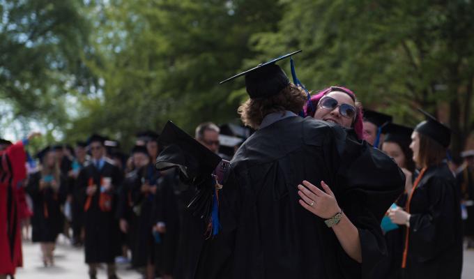 Two graduating students hugging each other in front of line of graduates in gowns
