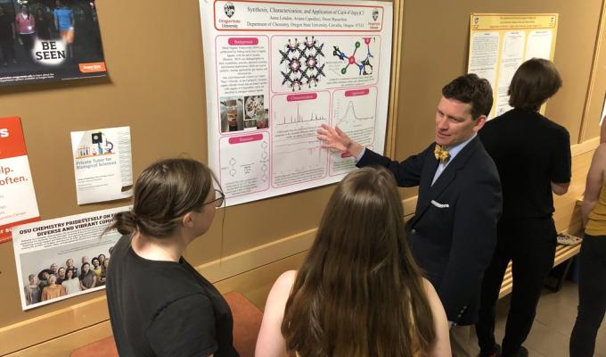A student shows a group of people a research poster hanging on a wall. 