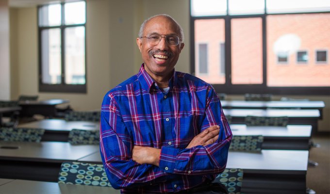 Portrait of Dr. Nathanial Whitaker: An African American male professor sitting in front of his classroom, folding his arms and smiling. 