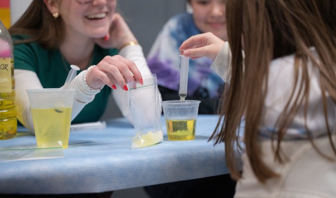 An OSU student smiles widely at the child in front of them. She holds a baggie of yellow liquid that the child is using a syringe to extract and place into a cup in front of her.