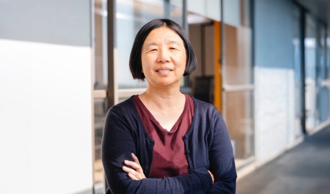 Headshot of Professor Bin Yu of UC Berkeley: woman in dark blue cardigan and maroon blouse, smiling with arms crossed while standing in front of college building with large glass windows.