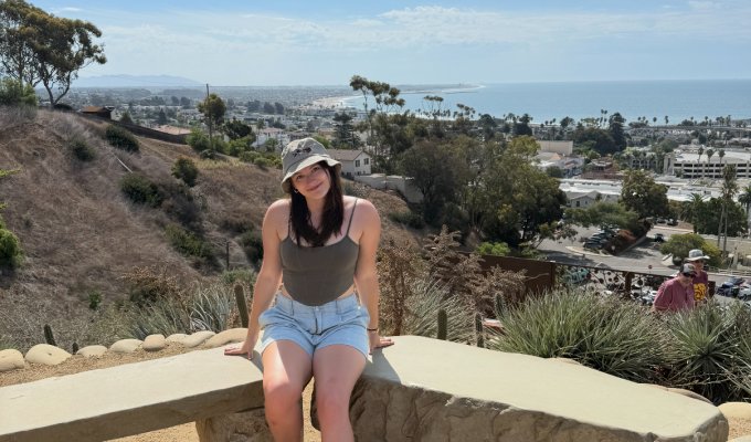 A woman in shorts and a hat poses for a photo near the ocean.