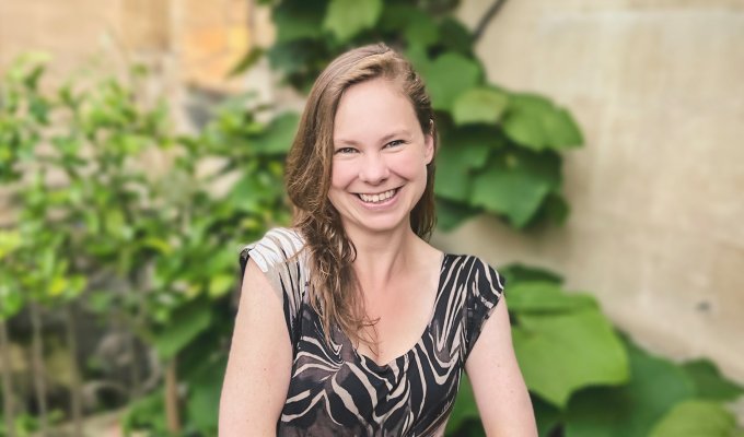 Headshot of Dr. Laura P. Schaposknik: A woman smiling with her hands folded in front of her, sitting in front of a background featuring beige stone walls and green plants.