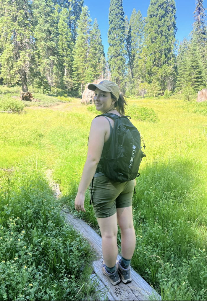 A woman stands on a log outside wearing a tank top and shorts.