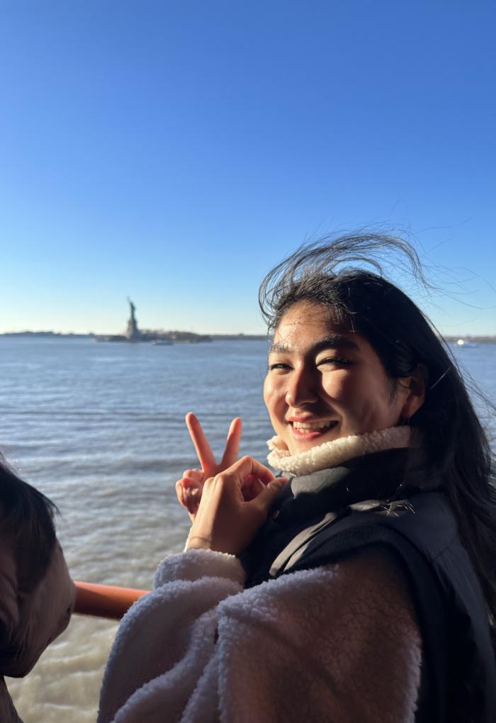 The wind blows through a woman's hair as she holds up a peace sign in front of a distant Statue of Liberty.