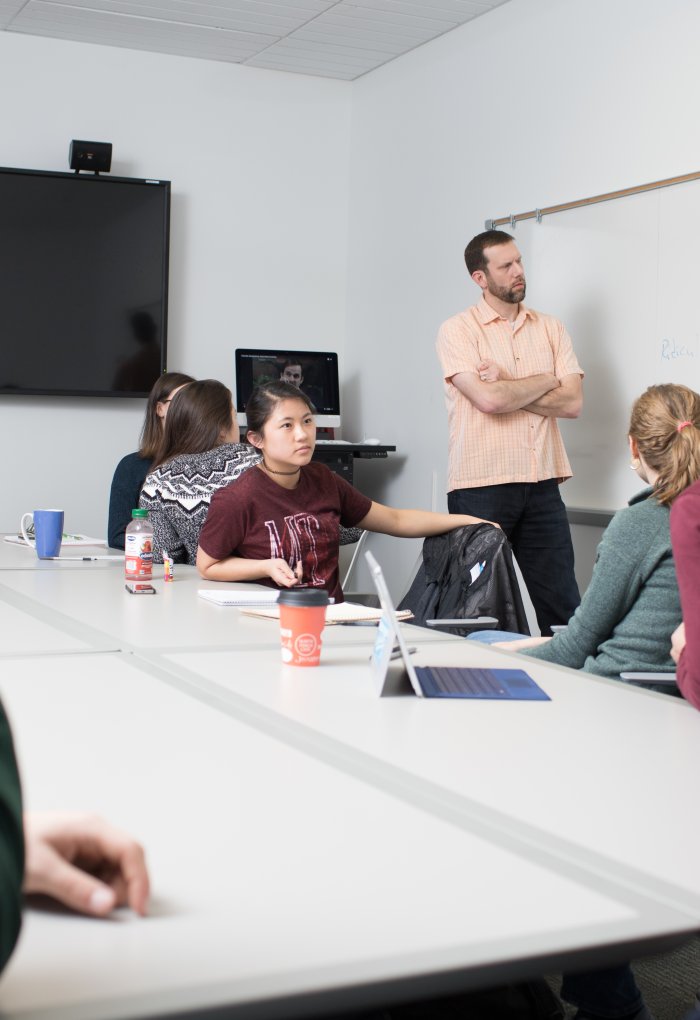 Students sit around a table and look towards an instructor in a salmon colored shirt.